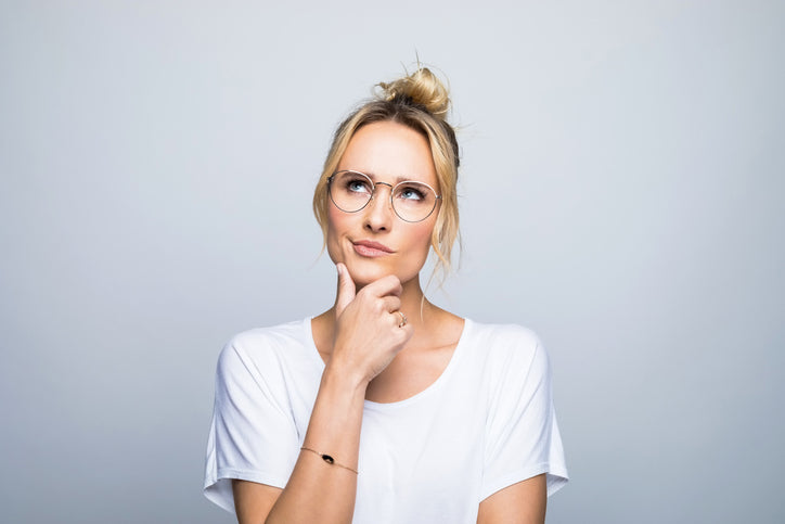 Woman with glasses resting her hand on her chin, looking up thoughtfully as she decides whether to use LED before or after microneedling